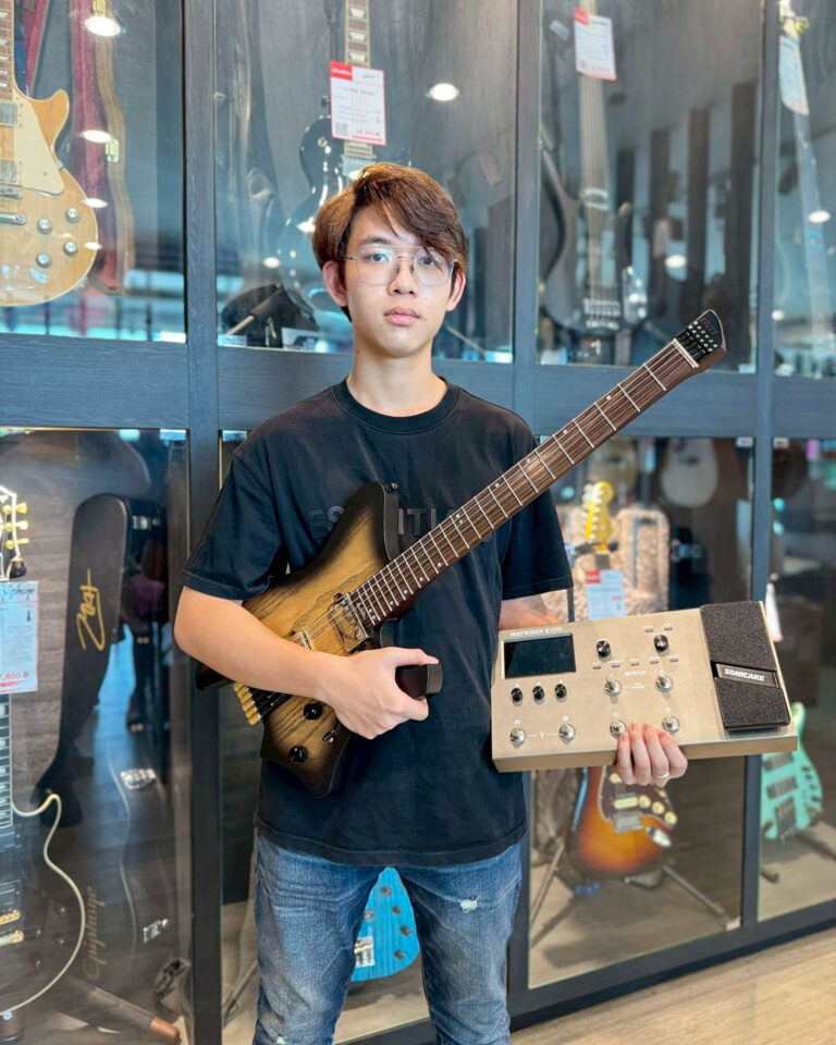 Young man in a black shirt stands in a guitar shop, holding an electric guitar and a pedalboard-like device in front of glass display cases with guitars.