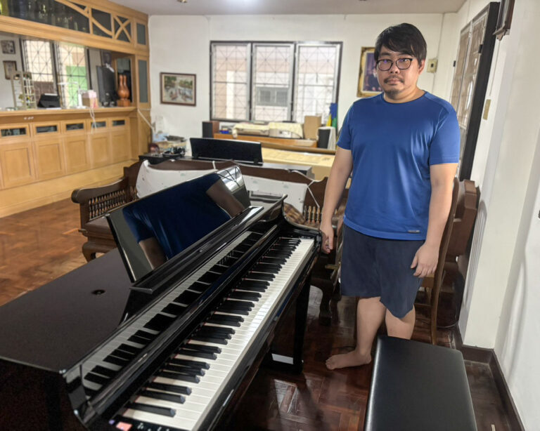 Man in a blue athletic shirt and glasses stands barefoot beside a glossy grand piano in a warmly furnished room with wooden cabinets and chairs nearby.
