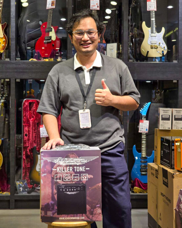 Smiling store employee giving a thumbs up while holding an Ashdown Killer Tone amplifier box in a guitar shop with guitars on display behind him