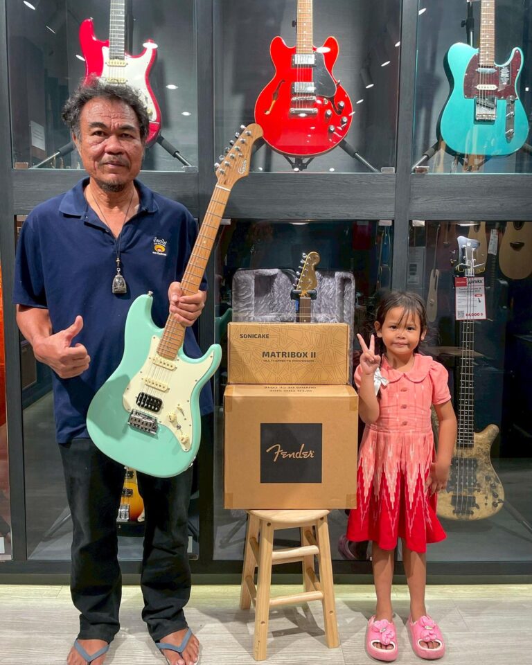 Man stands with a mint-green Fender guitar while a young girl in a pink dress poses beside stacked boxes in a guitar shop display behind them.