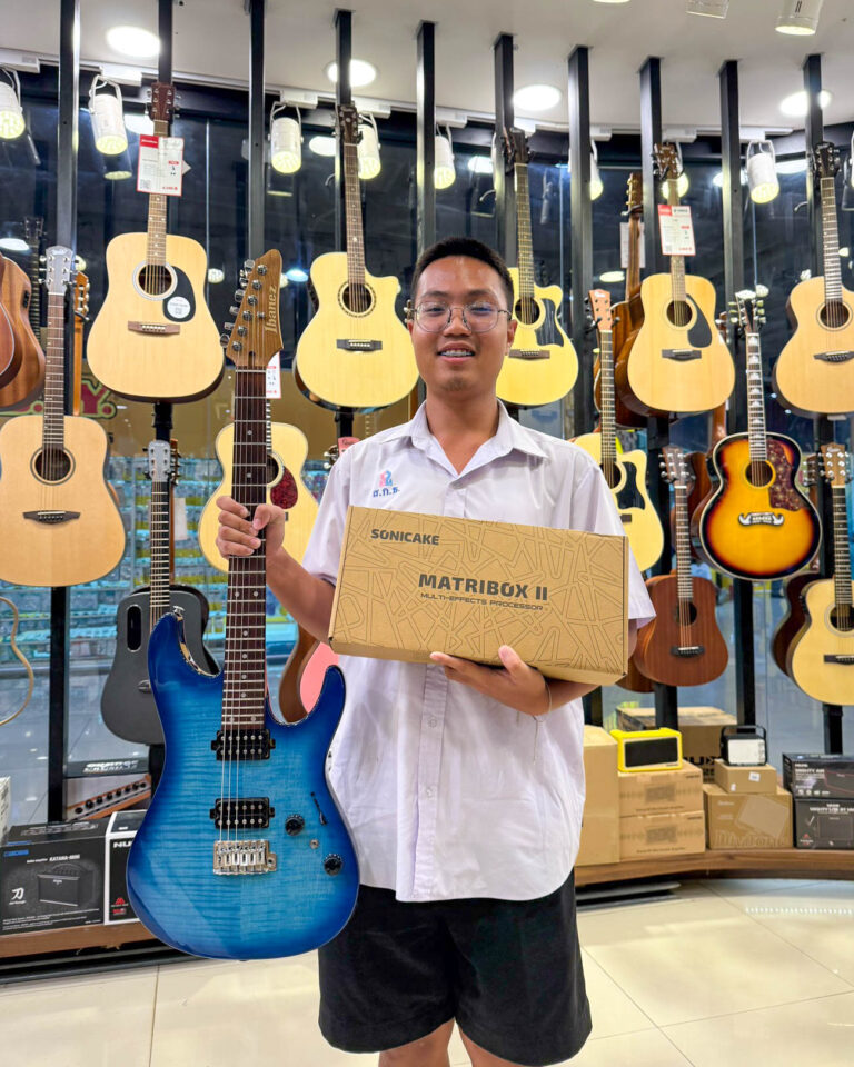 Man in a white shirt stands in a guitar shop, smiling, holding a blue electric guitar in one hand and a boxed multi-effects processor in the other, with guitars on display behind him.