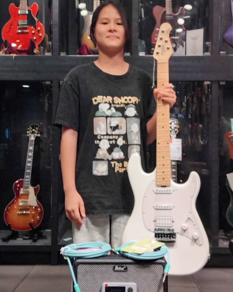 Person standing in a guitar shop, holding a white Stratocaster-style electric guitar with a small turquoise amplifier and coiled cables in front.