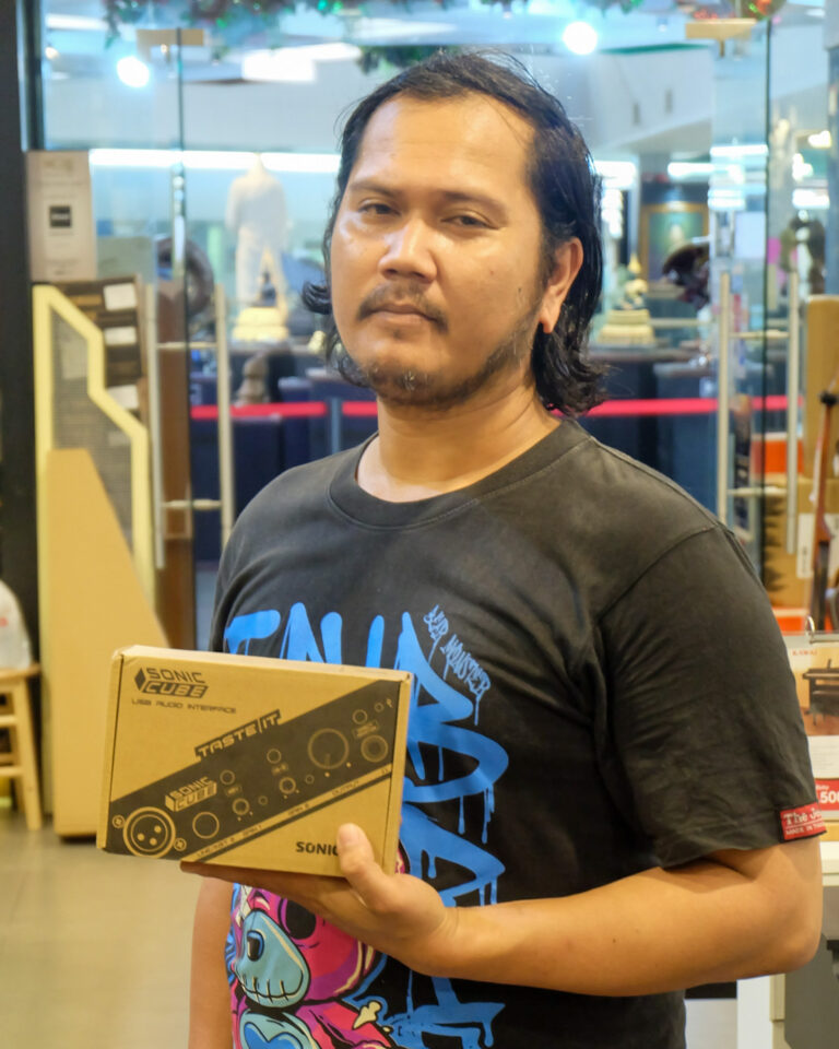Man in a black graphic T-shirt holding a box labeled Sonic Cube USB audio interface, standing in a store.