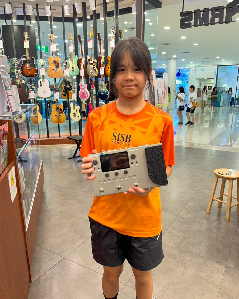 Young girl in an orange SISB Schools shirt holds a guitar multi-effects processor in a guitar shop with colorful guitars on display behind her.