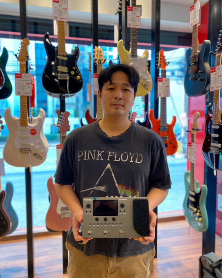Man stands in a guitar shop holding a guitar multi-effects processor in front of a wall of electric guitars.