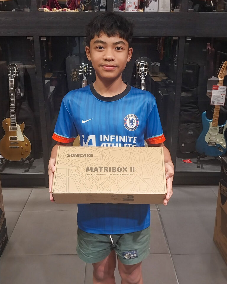 Boy wearing a blue Chelsea football shirt holds a Sonicake Matribox II box in a music shop, with guitars on display behind him.