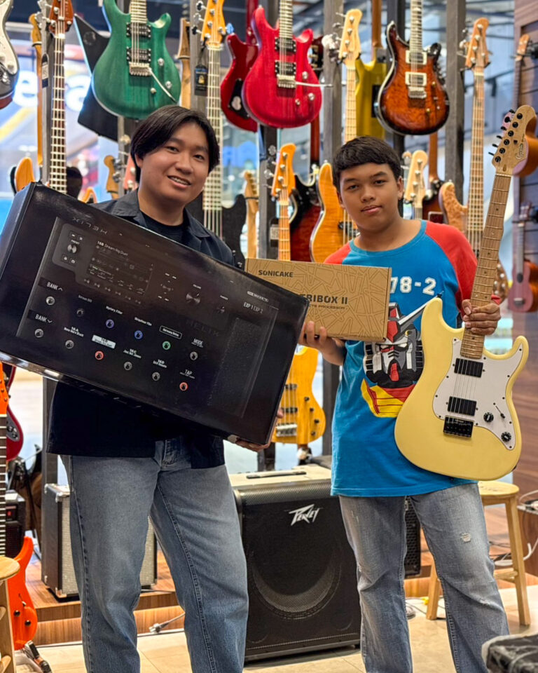 Two people stand in a guitar shop posing with new gear: a large black audio device and a boxed item, with many colorful guitars hanging behind them.