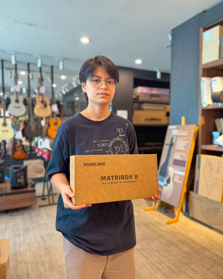 Person in a music store holds a boxed product labeled 'MATRIBOX II' in front of them; guitars hang in the background.