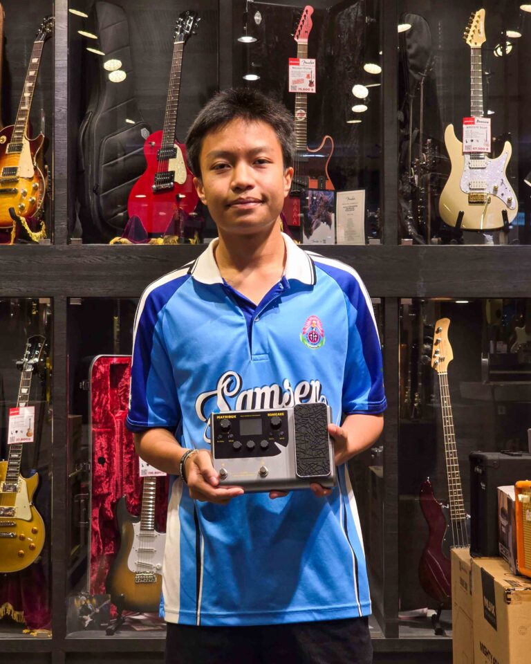 Person in a blue jersey stands in a guitar shop, holding a guitar effects pedal with display cases of guitars behind them.