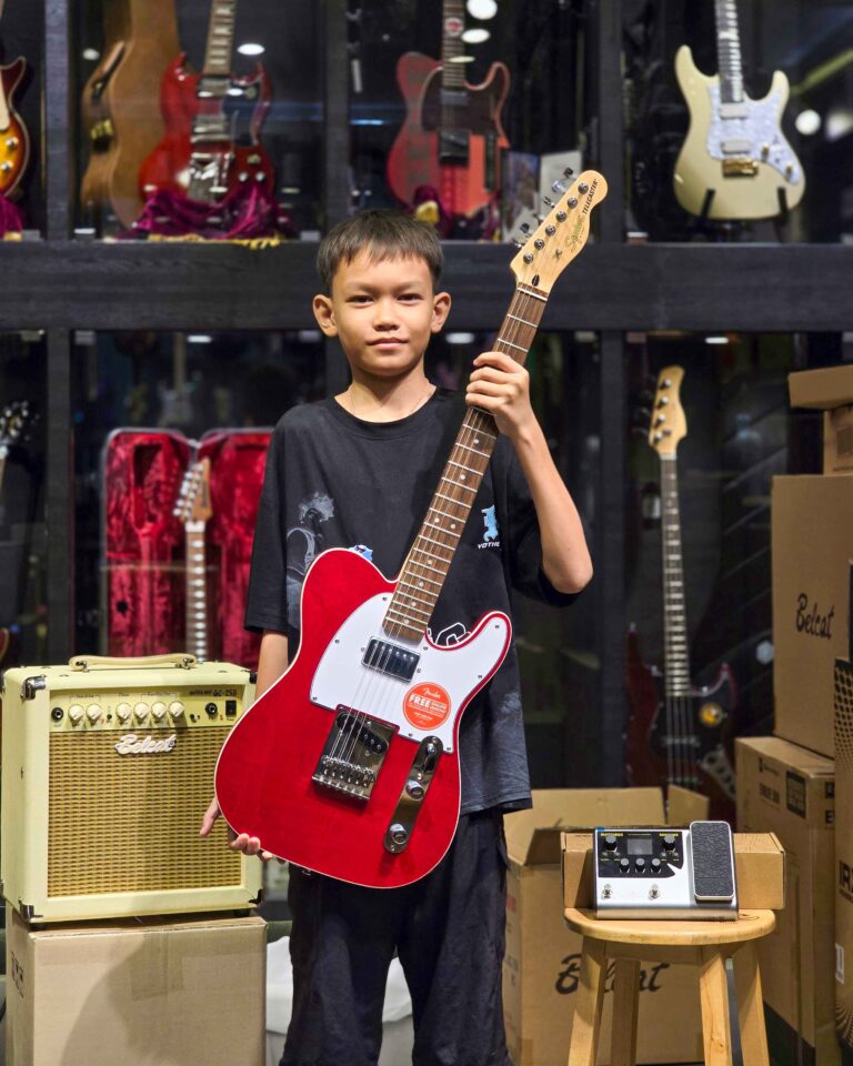 Boy holding a red electric guitar in a music store with amps and other guitars in the background.