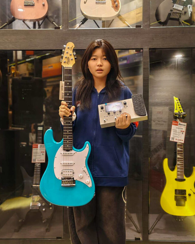 Young person in a blue hoodie holds a turquoise electric guitar with a white pickguard and a compact effects pedal in a music shop.