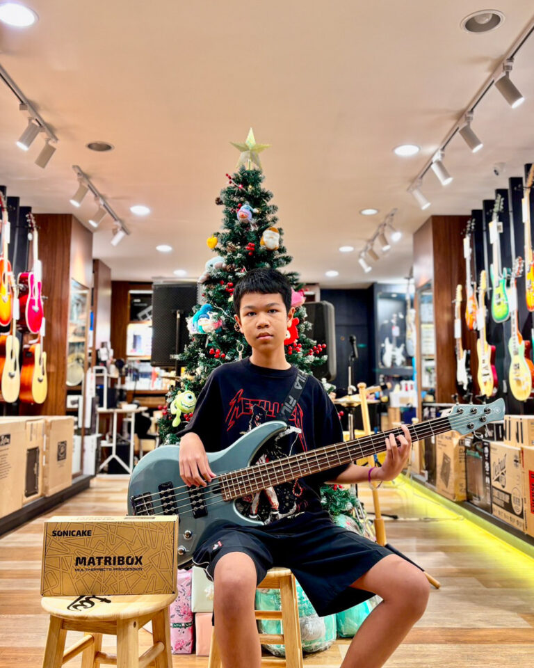 Boy sitting on a stool playing a teal bass guitar in a music shop, Christmas tree behind him with ornaments and guitars on the walls nearby.