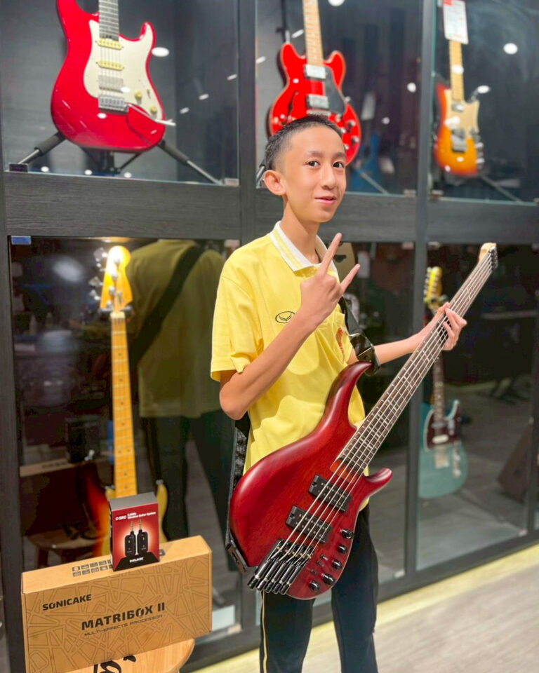 Young boy in a yellow shirt posing with a red bass guitar in a guitar shop display cases behind him.