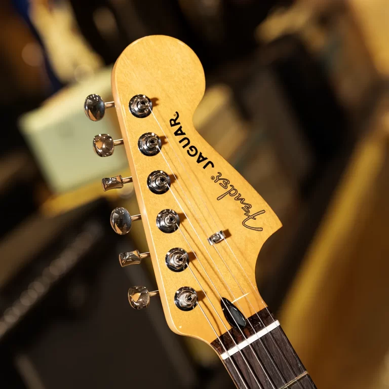 Close-up of a Fender Jaguar guitar headstock with six chrome tuning pegs, strings, and the Fender logo visible ขายราคาพิเศษ