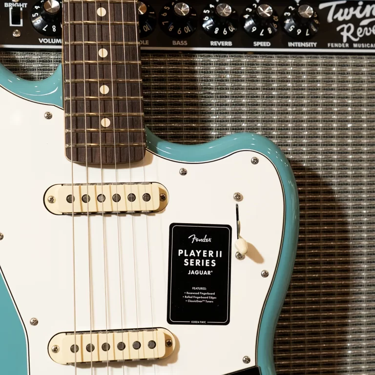 Teal Fender Jaguar electric guitar with white pickguard and gold pickups in front of a black amplifier panel labeled Master controls (focus on guitar). ขายราคาพิเศษ