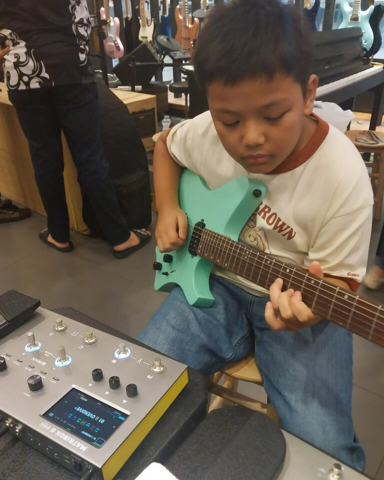 Young boy sits and plays a mint-green electric guitar in a guitar shop, with pedalboard in the foreground and guitars on the wall behind him.