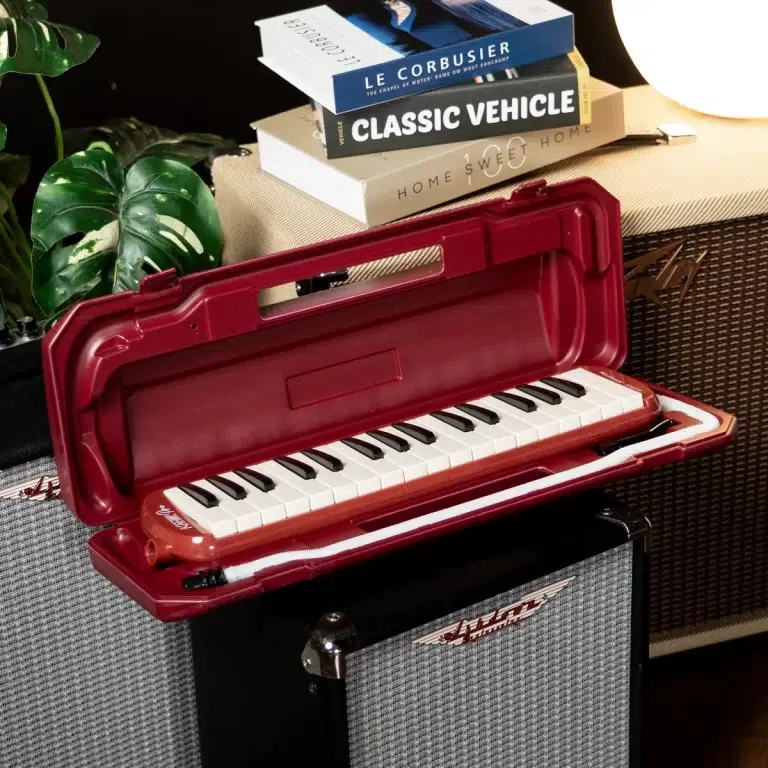 Red plastic melodica in an open case resting on a speaker amp with a stack of books and a plant nearby, in a cozy music corner. ขายราคาพิเศษ