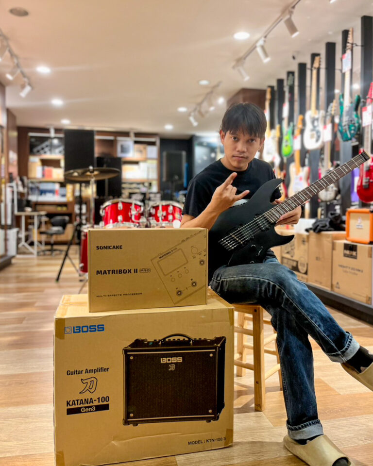 Young man in a guitar shop sits on a stool with a black electric guitar, next to boxed Boss amplifiers on the floor.