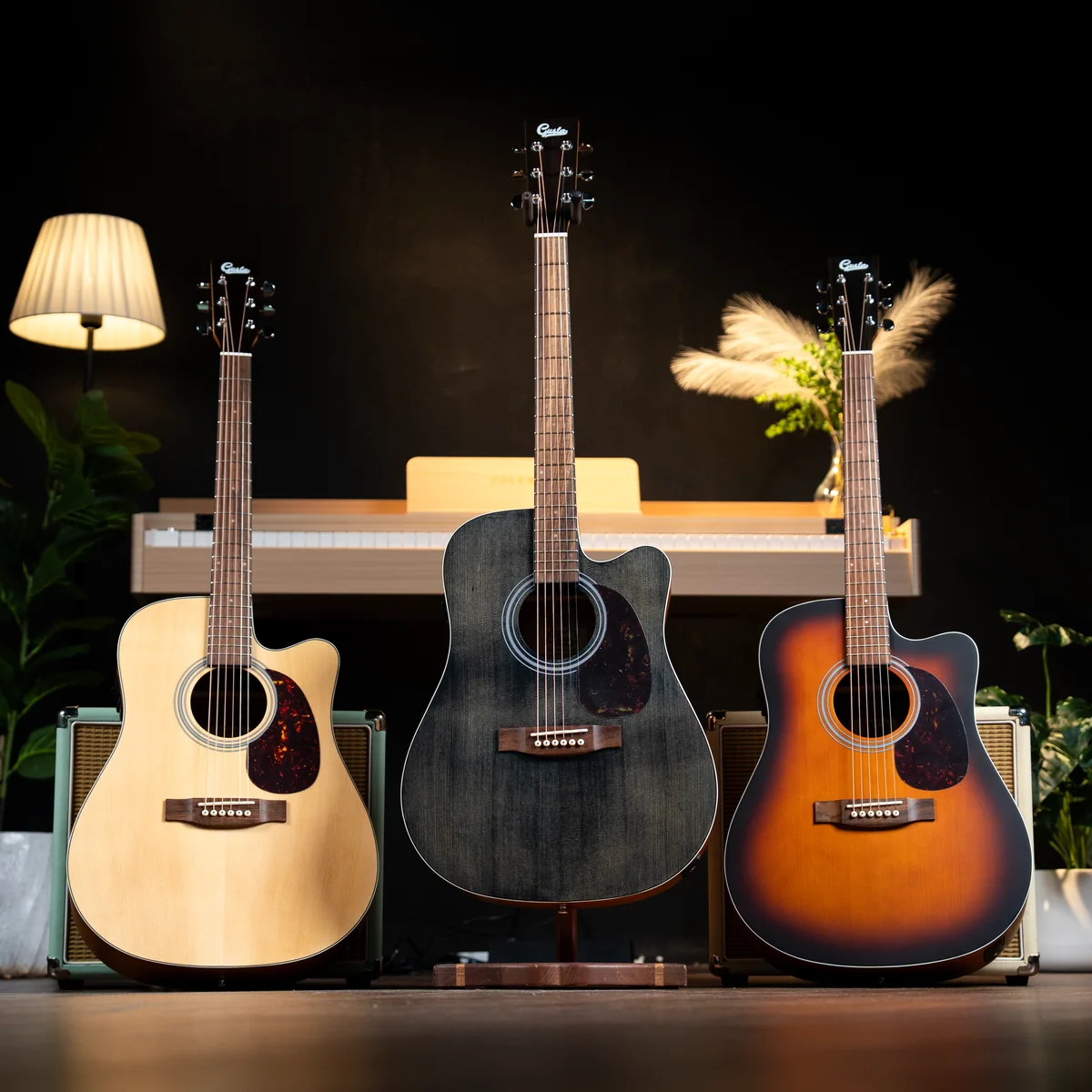Three acoustic guitars on stands in a music setting, left natural, center black, right sunburst, with a piano and amps behind them.
