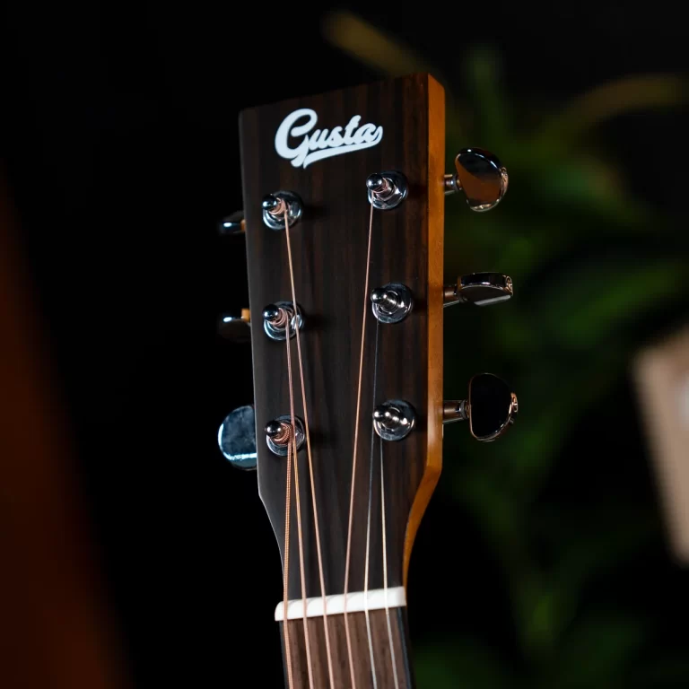 Close-up of a guitar headstock showing six tuning pegs, strings, and the 'Gusta' logo on dark wood surface. ขายราคาพิเศษ