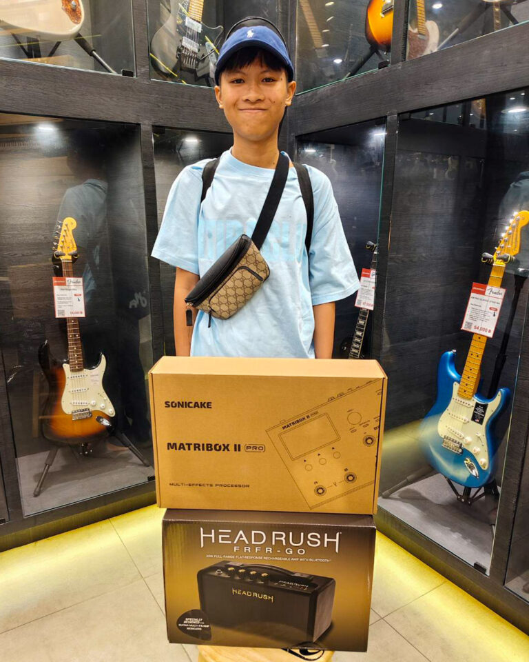 A smiling young person in a blue shirt and cap stands behind a stack of guitar gear boxes in a music store, with guitars on display behind glass.