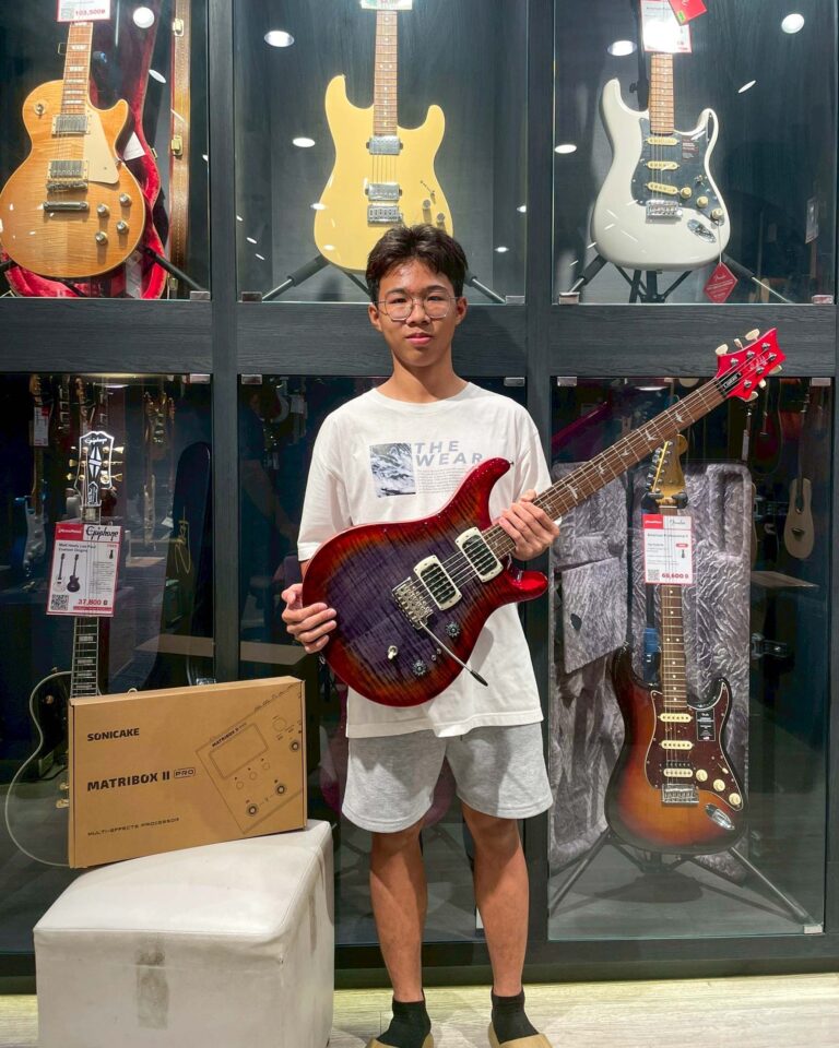 Young boy in a white T-shirt and glasses holding a red electric guitar inside a guitar shop display case.