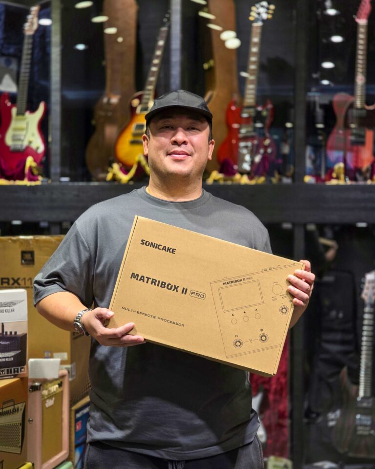 Man in a cap smiles while holding a large cardboard box in a guitar shop, with guitars on display behind him