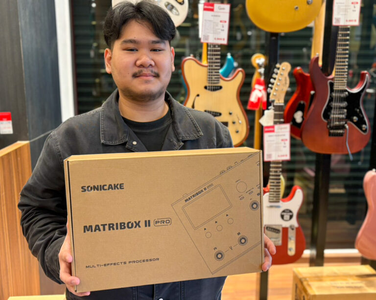 Man in a dark jacket holds a large cardboard box for a Sonicake MatriBox II Pro in a guitar shop, with guitars on display in the background.