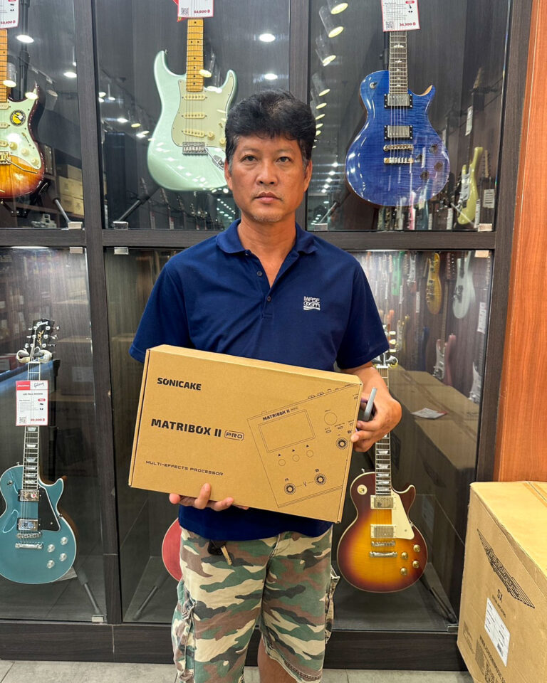 Man in a navy polo stands in a guitar shop, holding a boxed SonicaKe Matribox II Pro multi-effects processor with guitars on display behind him.