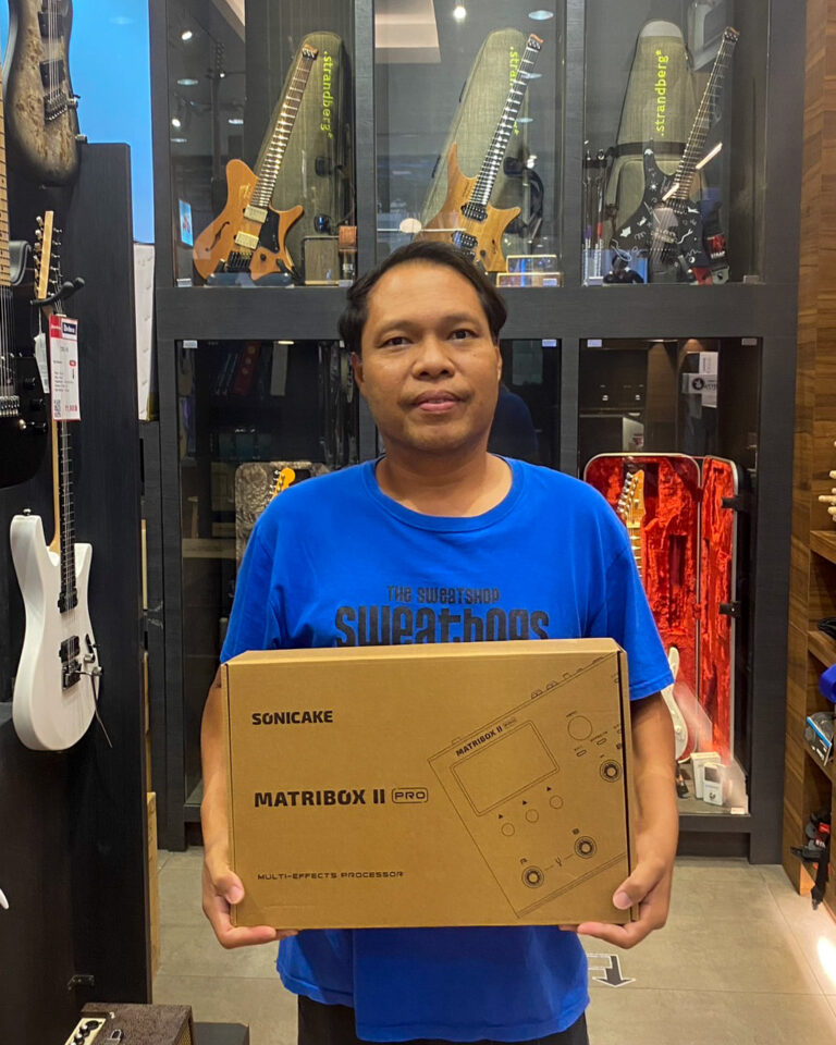 Man in a blue shirt holding a Sonicake Matribox II Pro box in a guitar shop, with guitars on display behind him.