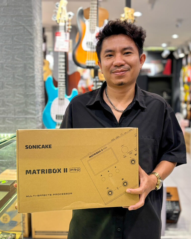 Man in a black shirt holding a cardboard box labeled Matibox II Pro multi-effects processor in a guitar shop.
