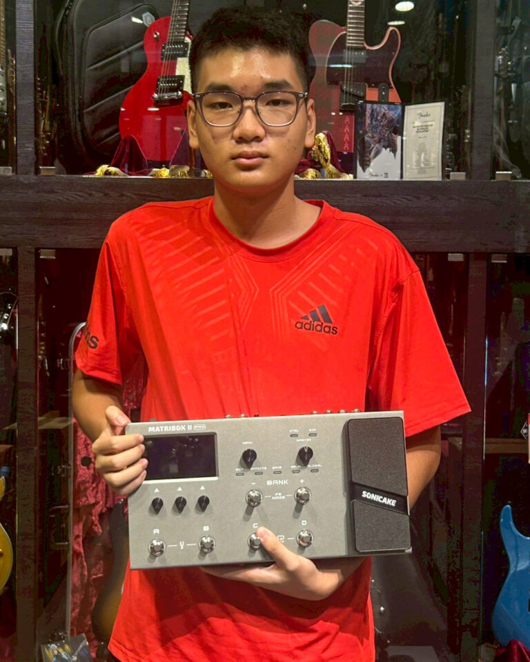 Young man in a red Adidas shirt holds a silver music production controller in a guitar shop, with guitars on display behind him.