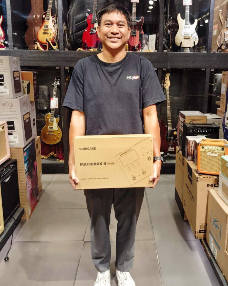 Smiling man in a black T-shirt holds a Sonicake MatrixBox II Pro box in a guitar shop with guitars and amps behind him.