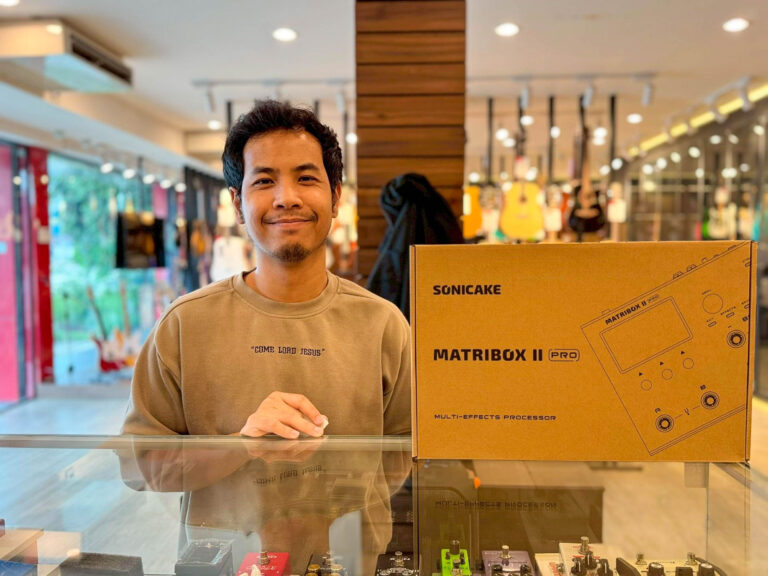 Young man in a store beside a large cardboard box labeled 'MATRIXBOX II Pro' holding a small item, with guitars on display in the background.