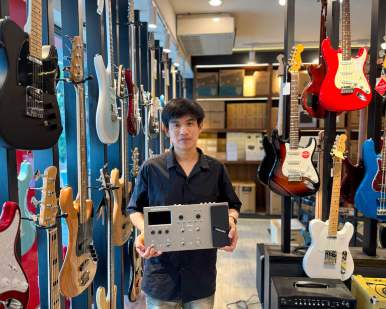 Man standing in a guitar shop, holding a rack-mounted audio device with knobs and buttons, surrounded by hanging electric guitars on blue display panels.