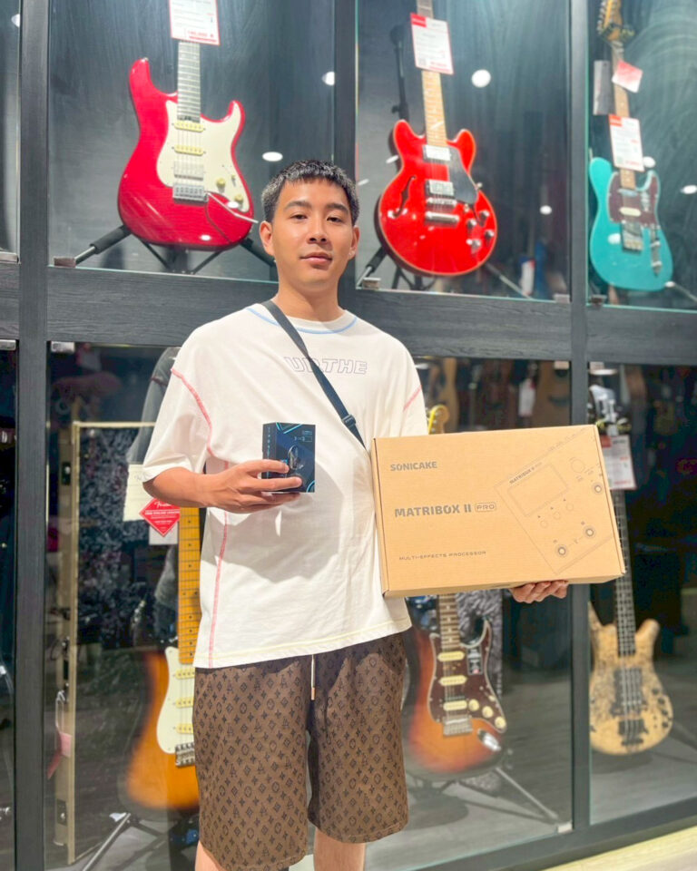 Man standing in front of a guitar shop display, holding a brown box labeled 'SONICAKE MATRIBOX II PRO' and a small blue box in his other hand.