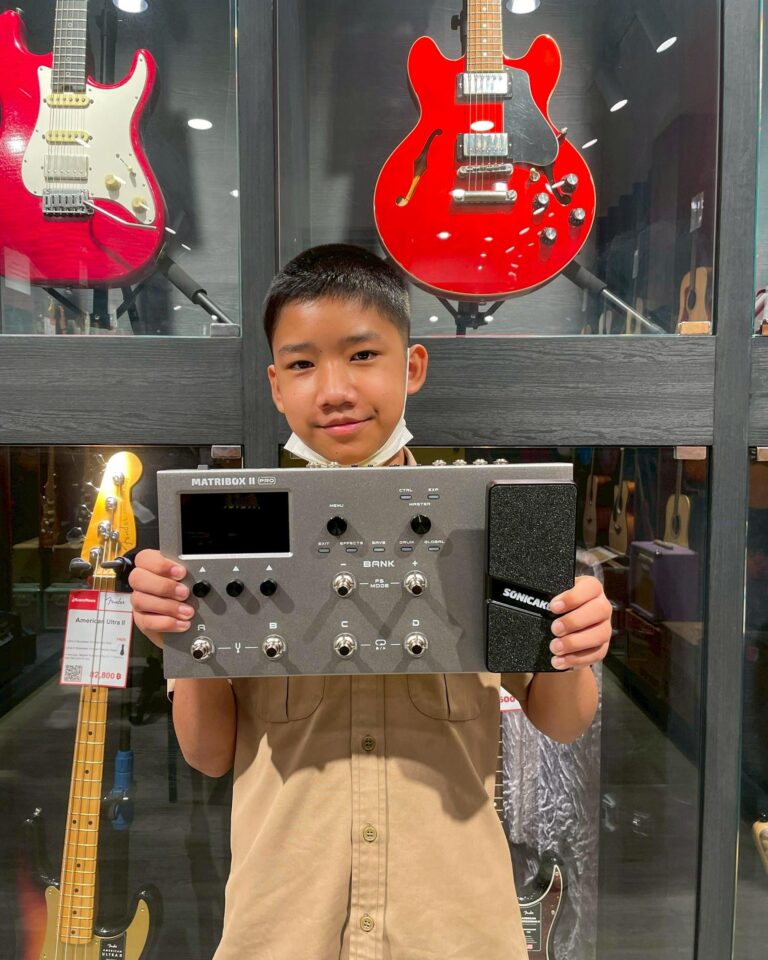 Smiling boy in a guitar shop holding a Matrixbox Pro music controller with red guitars on display behind glass.