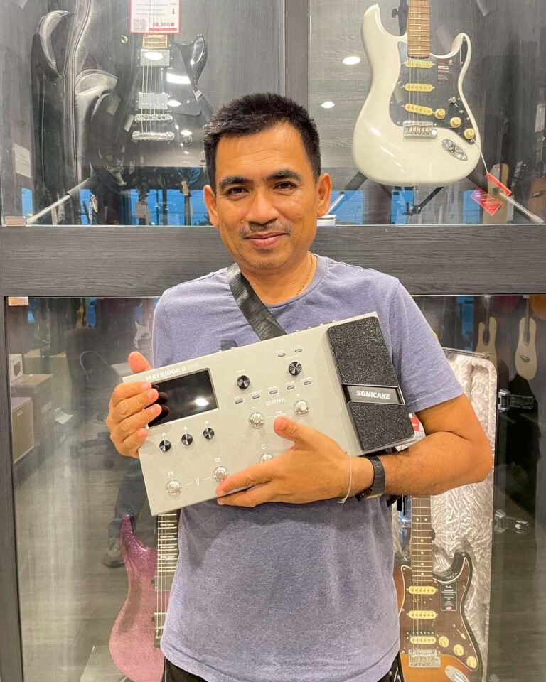 Man in a blue shirt smiles while holding a music production controller and a Sonicake pedal inside a guitar shop, with guitars on display behind him.