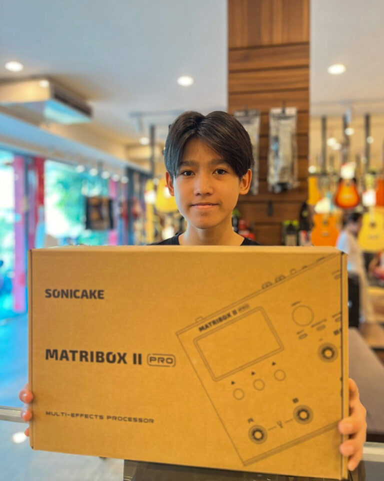 A young boy stands in a music store, holding a large cardboard box labeled Sonicake MatriBox II Pro multi-effects processor.
