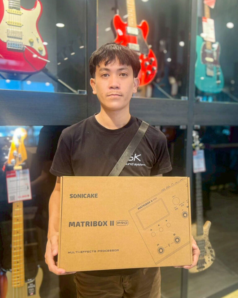 Person in a black shirt holding a boxed MatriBox II Pro multi-effects processor inside a guitar shop with displays of electric guitars behind.