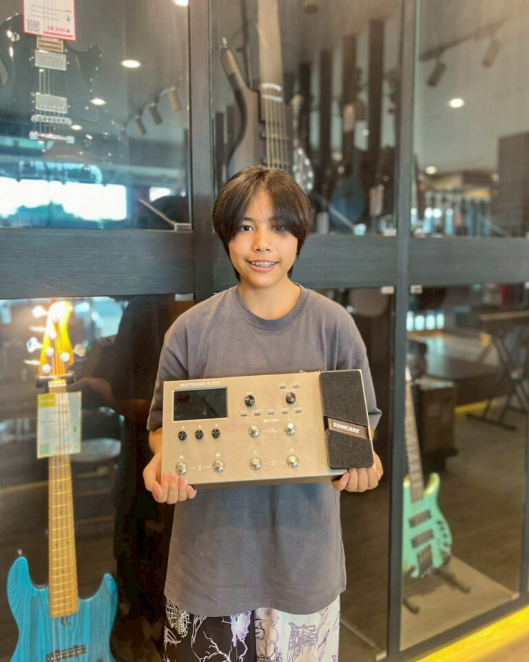 Smiling boy holds a metal guitar effects unit in a music store, with electric guitars on display behind him.