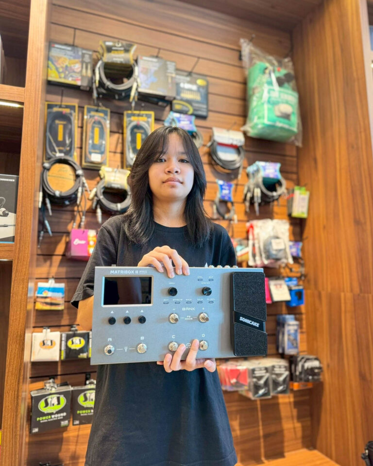 Young woman in a music shop holding a large blue music controller/display unit in front of a wall of gear.