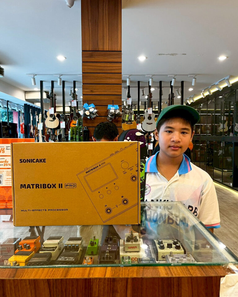Boy in a green cap stands beside a large SONICAKE MATRIBOX II PRO box at a guitar shop; guitars hang in the background.
