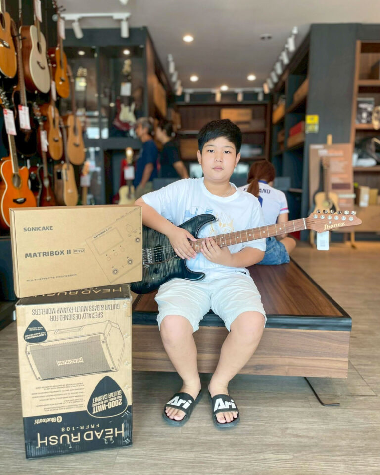 Young boy in a music store sits on steps, holding a black electric guitar with guitar boxes nearby and guitars on display behind him.