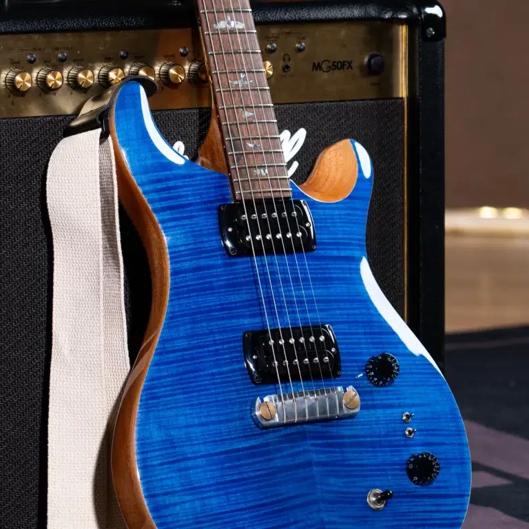 Blue electric guitar with a flame maple top leaning against a black amplifier, cream strap draped over the side, chrome hardware and knobs visible. ขายราคาพิเศษ