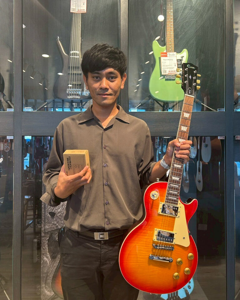 Man standing in a guitar shop, holding a sunburst electric guitar in his right hand and a small boxed item in his left.