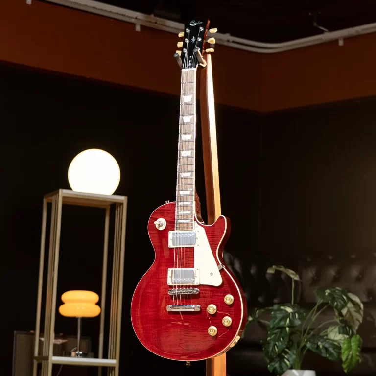 Red Les Paul–style electric guitar hanging on a stand in a dimly lit room, with a glowing round lamp and a leafy plant nearby ขายราคาพิเศษ