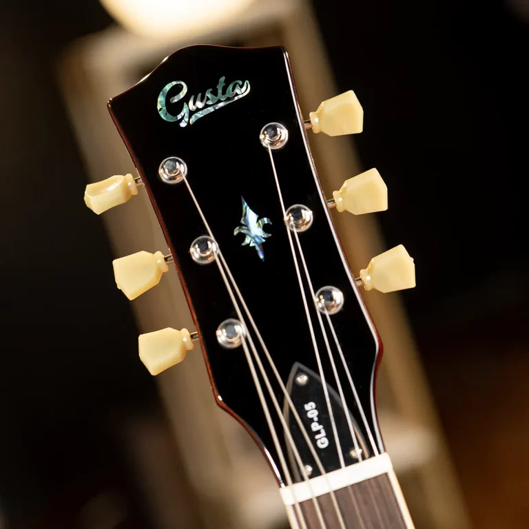 Close-up of a black guitar headstock with cream tuning pegs and silver strings; brand logo 'Gusta' visible with a decorative inlay. ขายราคาพิเศษ