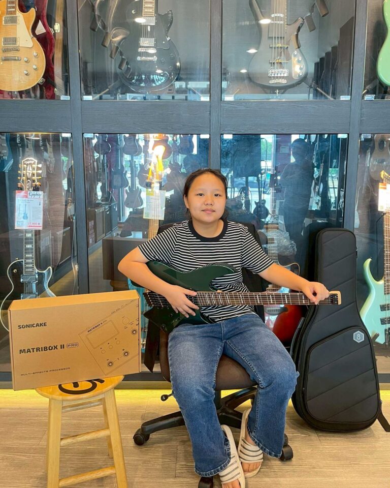 Girl sitting in a guitar shop, holding a green electric guitar with guitars on display behind glass cases and a cardboard box nearby.