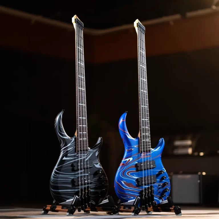 Two electric bass guitars with marbled finishes (black/gray and blue/red) standing on black stands against a dark stage backdrop, ready for display or performance. ขายราคาพิเศษ